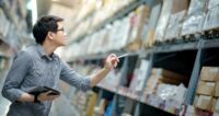 young Asian Man Worker Doing Stocktaking Of Product In Cardboard Box On Shelves In Warehouse By Using Digital Tablet And Pen. Physical Inventory Count Concept